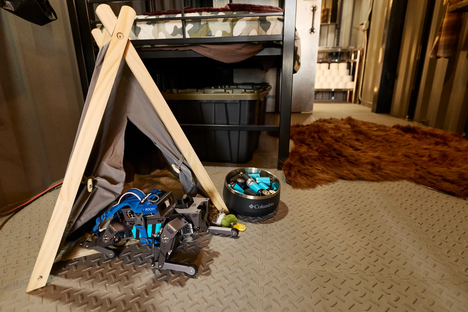 Sığınakta küçük çadırın yanında duran Rocky adlı robot köpek ve pil dolu kabı.