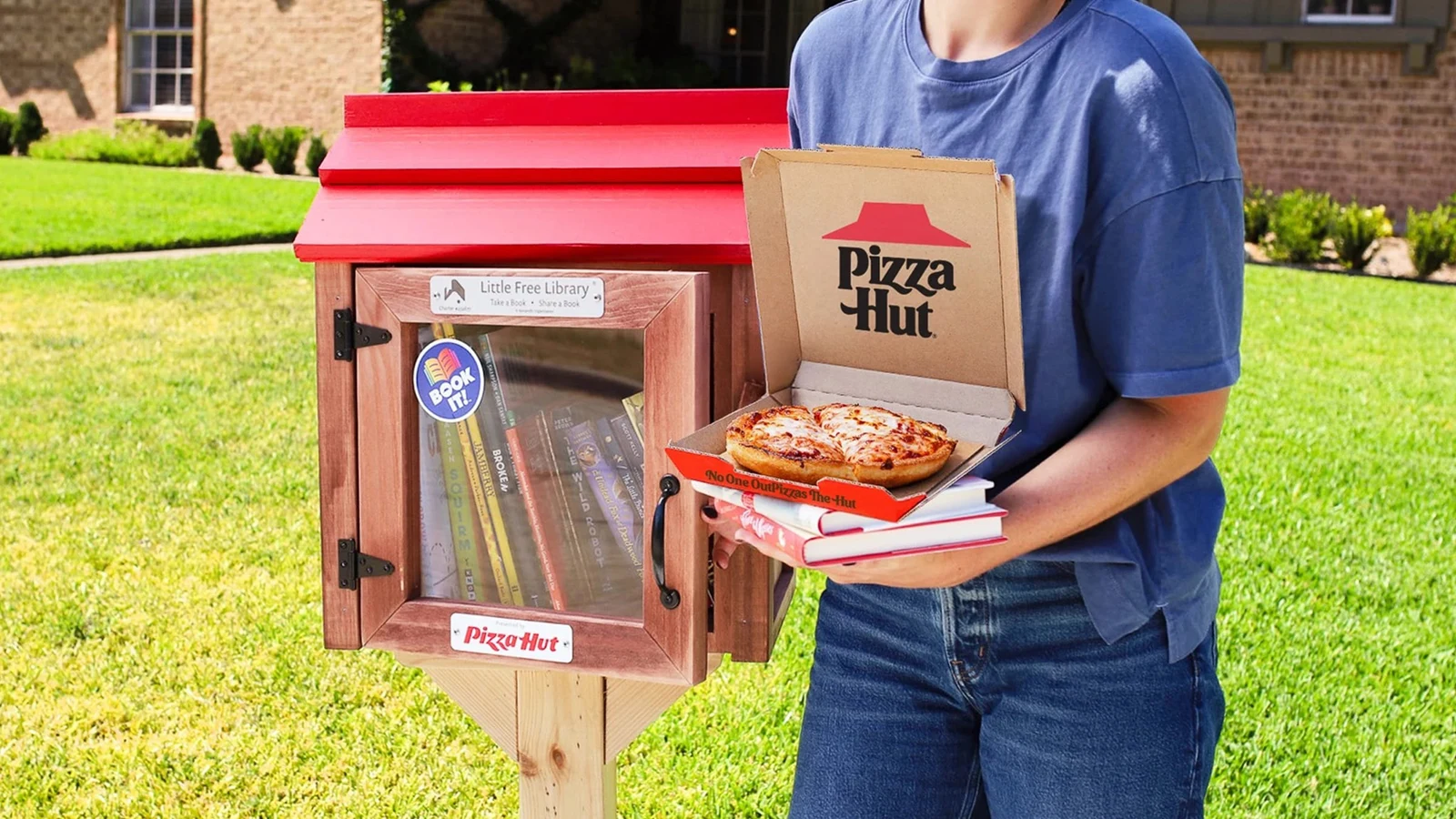 Kırmızı çatılı mini kütüphane yanında kitap ve pizza taşıyan kişi