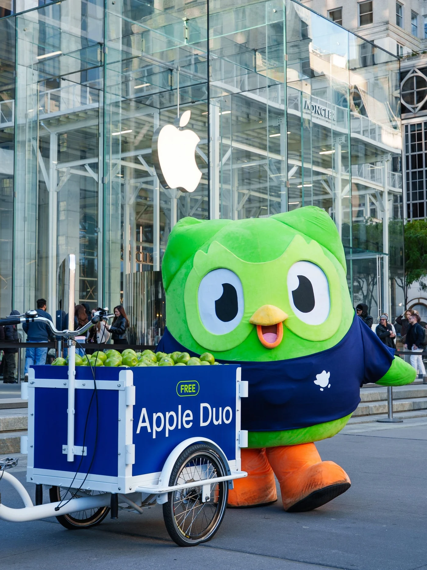 Apple Fifth Avenue mağazasının önünde lacivert tişörtlü Duo ve Apple Duo yazılı yeşil elma arabası.